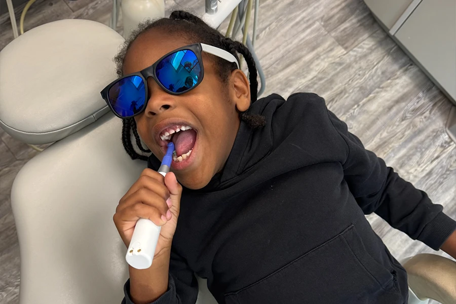 Child wearing blue mirrored sunglasses brushing teeth in dental chair