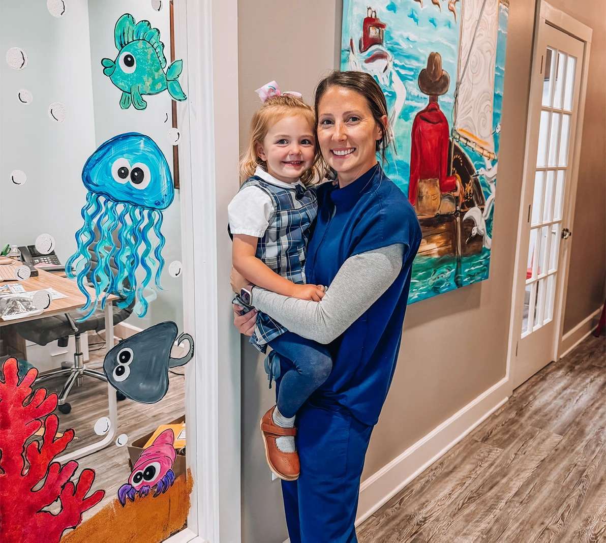 Dental staff member in blue scrubs holding a smiling young child in a colorful ocean-themed dental office hallway